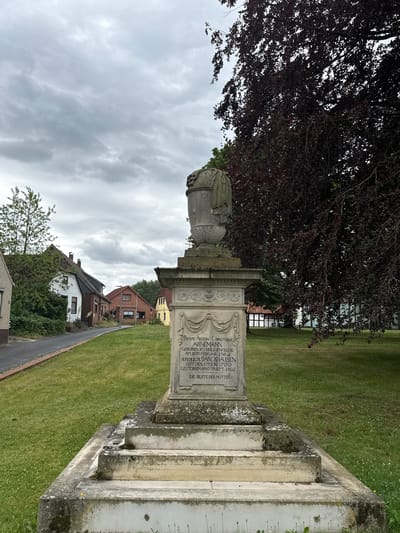 This is a vertical, eye-level, medium shot of a monument with text inscribed on it, set against a backdrop of a cloudy sky, a large tree with dark foliage, and a grassy area with some houses and trees in the distance. The monument appears to be made of stone, with a carved urn-like feature at the top. The inscription on the monument is in German and appears to be a dedication or memorial. The text includes names and dates, suggesting it is a gravestone or memorial plaque. The overall mood of the image is somber, possibly due to the overcast weather and the nature of the monument. The location appears to be a public space, likely a cemetery or a park, given the presence of the monument and the surrounding greenery. The houses in the background indicate a residential or village setting. The image was likely taken during the daytime, given the visibility of the scene.