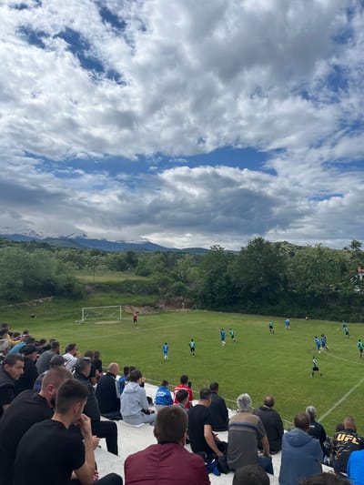The image shows a soccer game viewed from stadium seating. A crowd watches players on a green field; one goal is visible. Mountains are visible in the background. The sky is filled with cumulus clouds. A person in a UFC shirt is in the crowd. The location is likely Kornitsa, Bulgaria.