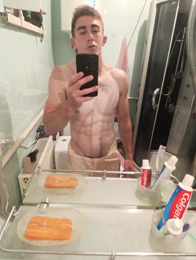This media file shows a young man taking a mirror selfie in a bathroom. He is shirtless and wearing a towel around his waist. He has a lean, muscular build. In the mirror's reflection, we see a shower enclosure, a toilet, and a radiator. The bathroom appears to be in a residential setting, likely in Fastiv, Ukraine, given the location context. On the shelf below the mirror are two bars of orange soap and some toiletries, including tubes of Colgate toothpaste and toothbrushes. The lighting suggests it might be daytime or early evening. The man's expression is neutral. There is visible text on the toothpaste tubes which reads "Colgate".