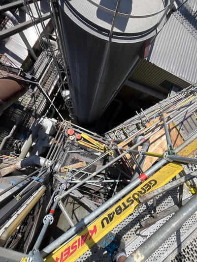 The image captures an overhead perspective of an industrial setting, likely a construction or maintenance site, viewed from a high vantage point. Scaffolding dominates the scene, constructed from metallic poles and platforms, suggesting ongoing work. Prominently displayed on yellow planks are the words "GERUSTBAU" and "KAISER," indicating the brand or type of scaffolding and potentially a company name.
A large, cylindrical metallic structure, possibly a silo or tank, curves into the frame from the left, its textured surface reflecting sunlight. A corrugated metal roof and a portion of a building with horizontal siding are visible in the upper left quadrant. Numerous pipes, conduits, and industrial equipment are interspersed throughout the scaffolding and structures, hinting at the functional purpose of the location.
The lighting suggests a bright, sunny day, casting sharp shadows across the metallic surfaces and scaffolding. The angle of the shot, looking down, creates a sense of depth and height. No people are clearly visible, though a person's legs and footwear are partially seen in the lower right corner, suggesting their presence within the scaffolding structure. The overall impression is one of industrial activity and construction, with a focus on the complex network of scaffolding. The location context of Neuss, Germany, suggests this is likely a port or industrial area.