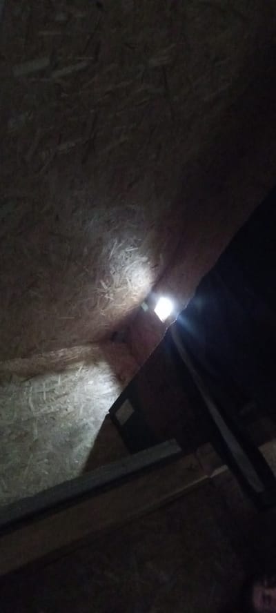 The image shows the interior of a dimly lit space, possibly an attic or a shed, with walls and ceiling constructed from particle board. A small, square opening in the ceiling allows a bright light source, possibly daylight, to penetrate the space. In the foreground, there are wooden beams and a darker object, possibly furniture or equipment, casting shadows. The overall impression is one of a neglected or unfinished interior space. The image location is identified as Заречный ауылдық округі, Kazakhstan. There are no people clearly visible in the image, although a faint suggestion of a face may be present in the lower right corner. The scene is static, with no apparent action or interaction taking place.