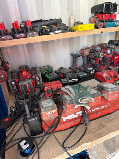 This image depicts a shelf and a table laden with various power tools and accessories. On the top shelf, there are numerous red and black power tool batteries, some with "REDLITHIUM M12" markings, and other battery packs. Towards the right, there are black and red power tools, including a drill and what appears to be a charger. A yellow box labeled "Klingspor Schleiftechnologie" is also visible.
The lower section shows a collection of power tool batteries, including several red Milwaukee brand batteries with "5.0" and "6.0" capacities, and some green batteries. There are also red power tools, one with a "HILTI" logo and "C 4/36-MC4" marking. A Motorola walkie-talkie, a power strip with a red switch, a spool of blue wire, and a pair of green work gloves are also present. The overall environment appears to be an interior space, possibly a workshop or storage area, with unfinished wooden surfaces and corrugated metal walls. The image does not contain any people or indicate the specific location within Janowice, Poland.