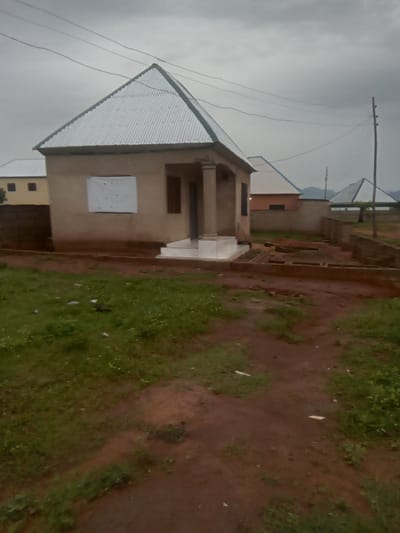 The image shows a small, single-story building with a corrugated metal roof, located in Zanzi, Nigeria. The building is painted a light tan color and has a covered porch supported by two pillars. A white sheet is covering a window on the front of the building. The area around the building is unpaved, with dirt paths and patches of grass. In the background, other buildings with similar roofs are visible, as well as a power pole and electrical wires. The sky is overcast, suggesting a cloudy day. There are no people visible in the image. The scene appears to be in a residential or semi-rural area.