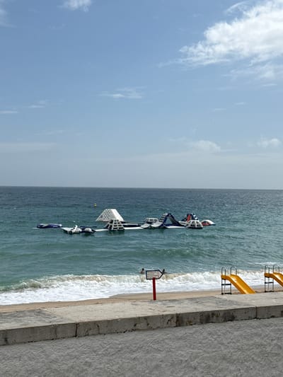 This is an image of an inflatable water park situated in the ocean near a beach. The water park features several inflatable structures with white and blue accents, including slides and climbing walls. Some people are visible in the water near the park, and one person is on the beach. Yellow slides are also present on the shore, leading down towards the water. The scene is set on a bright day with a clear blue sky and scattered white clouds, suggesting fair weather. The ocean is a vibrant blue-green with moderate waves crashing on the sandy beach. A concrete walkway or seawall runs along the right side of the image, with a red signpost with some text visible. The location is identified as Sesimbra, Portugal. The overall mood is one of leisure and recreation on a summer day.