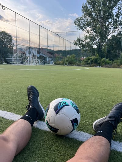 The image depicts a close-up view of a person's legs and feet, clad in black soccer cleats and black socks, positioned on an artificial turf soccer field. A soccer ball, featuring a white base with teal, black, and purple geometric patterns, rests on the field in front of them, slightly to the right of the center of the frame. The soccer field is lined with white markings, and a chain-link fence encloses the playing area. Beyond the fence, residential buildings and trees are visible under a partly cloudy sky. The scene appears to be an outdoor soccer facility.