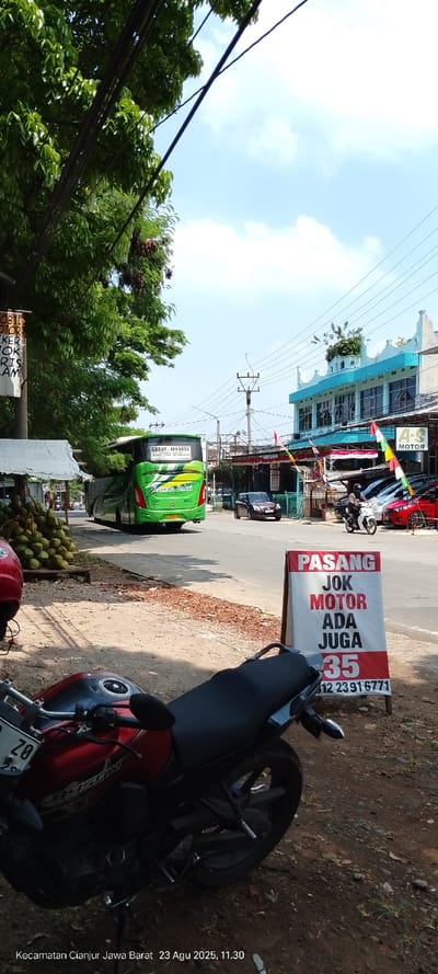 This image captures a daytime street scene in Kecamatan Cianjur, Jawa Barat, Indonesia, specifically along a road in Nagrak, on August 23, 2025, at 11:30 AM.
In the foreground, a red and black Yamaha Byson motorcycle with a partial license plate "Z 8" is parked on a dirt shoulder, facing right. To its right, a white A-frame sign with red lettering advertises "PASANG JOK MOTOR ADA JUGA 35" (Motorcycle seat installation also available 35) and a contact number (0812 2391 6771). The immediate left foreground is dominated by the dense green canopy of a tree, with multiple dark electrical wires running across the upper left quadrant of the frame.
The midground features a paved two-lane road with a visible center line. A large, green passenger bus, marked "GABUT - JAKARTA" and "KARENA BAKTI" with license plate "D 7536 AL," is moving away from the viewer on the road, heading right. Behind the bus, a dark-colored compact car follows. On the far right side of the road, a person wearing a dark helmet and jacket rides a white scooter, also moving away from the viewer.
Along the left side of the road, a makeshift stall with a corrugated metal roof is visible, with a large pile of green coconuts on a stand. Across the street, on the right side, a row of commercial buildings lines the road. A prominent two-story building features a distinctive blue and white facade with ornate rooftop architectural elements. Adjacent to it, another building with red awnings and numerous small colorful flags displays a sign for "A.S. MOTOR." Several vehicles, including a red car and a white scooter, are parked in front of these establishments. Multiple utility poles with overhead wires are visible along the street.
The background shows a clear, bright blue sky with scattered white clouds, indicating sunny weather.