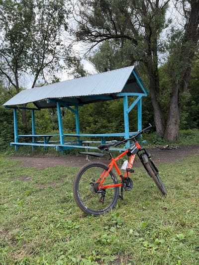 An orange bicycle is parked on a grassy area next to a blue picnic shelter in Subkhankulovo, Russia. The shelter has a corrugated metal roof and wooden benches. Tall trees with green foliage are visible in the background, along with a cloudy sky. The ground is covered with grass and dirt. A fire pit with ashes is present near the shelter. The bicycle is leaning on its kickstand and has a water bottle attached to the frame. The scene appears to be a park or recreational area.