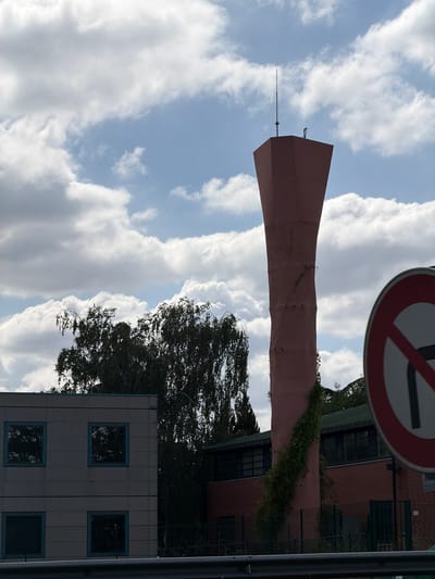 The image shows a tall, conical, pink structure, likely a water tower, in the city of Évry-Courcouronnes, France. The structure has a lightning rod at its apex and is partially covered in ivy. To the left of the water tower is a modern, grey building with several square windows. Behind the water tower and to its right is a red brick building with a green roof. A traffic sign, indicating a prohibition of turning, is visible in the foreground on the right side of the image. The sky is cloudy, with patches of blue visible. The overall scene appears to be an industrial or urban area. There are no people visible in the image.