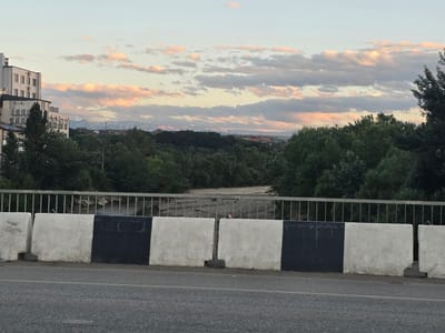 The image depicts a road scene in Khasavyurt, Russia, with a concrete barrier painted in alternating black and white sections in the foreground. Beyond the barrier, a metal railing lines the edge of what appears to be a bridge or elevated roadway. A dry riverbed or rocky creek bed is visible below the railing, bordered by dense green trees. In the distance, more trees and scattered buildings are visible, with mountains forming the backdrop under a sky filled with soft, pink and grey clouds, suggesting it is either sunrise or sunset. Several buildings, some appearing to be residential, are situated to the left of the frame, with one multi-story building featuring visible air conditioning units. The overall impression is a view from a bridge overlooking a natural landscape within or near the city.