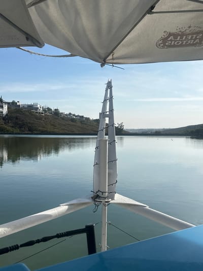 A tranquil view of a calm body of water under a pale blue sky. Reflections shimmer on the water's surface, mirroring the distant shore. A partially visible white sun umbrella with "Stella Artois" branding is in the foreground. A boat or structure with white rails and string lights further defines the foreground, adding a sense of being on the water.
