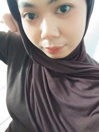A medium close-up selfie shows a young woman wearing a dark brown hijab and matching long-sleeved top. Her head is tilted slightly to the left, and her left hand is raised to her head, partially obscuring her ear. She is looking directly at the camera with a neutral expression. Her makeup includes subtle eye shadow and lipstick. The background appears to be an indoor setting with a white wall and some faint diagonal lines on the left side, possibly indicating a window or architectural feature. The lighting is soft, with some shadows visible on her face and clothing. The image was taken in Watampone, Indonesia.