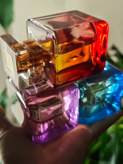 A hand is holding four stacked, colorful perfume bottles. The top bottle has amber and red liquid, the one below it is pink, and the bottom one is blue. The bottles are made of glass and have a faceted design. The background is blurred and appears to be an indoor setting with green foliage visible. There is no indication of the city of Kursk, Russia.