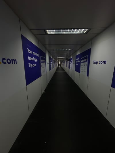An interior shot shows a dimly lit, long corridor with white walls displaying advertisements for Trip.com. The corridor appears to be under construction or renovation, with the white panels serving as temporary covering. Fluorescent lights are visible in the ceiling, providing minimal illumination. The floor is covered with a dark material, possibly carpet or rubber matting. The perspective is from the beginning of the corridor, looking towards a faint light at the far end, where a solitary figure is dimly visible. The advertisements are consistent on both sides of the corridor, featuring the Trip.com logo and promotional text in blue and white. The overall atmosphere is utilitarian and somewhat sterile.
