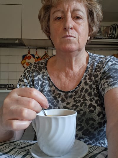 The media file shows a medium shot of a woman from the chest up, holding a white mug with a spoon inside. She is wearing a gray and black leopard-print t-shirt. The background appears to be a kitchen, with white tiled walls and cabinets visible. A checkered tablecloth is partially visible on the left side of the frame, and a decorative piece with fruit is hanging on the wall behind her. The lighting suggests it is daytime. The woman's expression is neutral. There is no visible text in the media file. The context provided indicates the location is Padua, Italy.