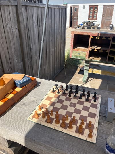 An outdoor scene in Skagen, Denmark, depicts a recreational area on a clear, sunny day. In the foreground, a light grey weathered wooden table supports a chessboard with light brown and dark brown Staunton-style pieces arranged as if in mid-game, with white pieces positioned on the lower half of the board. To the immediate left of the chessboard, a wooden box with internal dividers and a closed blue box are present. To the right, a rectangular silver-grey device, possibly a chess clock, and the base of a clear plastic bottle are visible. The midground features a tall, weathered vertical wooden fence running along the left side, against which a grey pole is secured with white ties. Adjacent to the table and fence, a distressed wooden cabinet, painted in multiple faded colors including red, green, yellow, and blue, stands on a light-colored paved surface. In the background, a white-painted building with two brown wooden doors and a matching window is visible. One door features a circular porthole-style window. A wooden picnic table with benches is positioned in front of the building. Strong shadows are cast across the scene, indicating direct sunlight. No individuals are present.