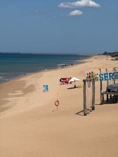 This is a horizontal, high-angle shot of a sandy beach and ocean in Tuy Hoa, Vietnam. The sky is a clear, bright blue with a few wispy white clouds. The ocean is a deep blue-green, with gentle waves breaking on the shore. A stone breakwater runs parallel to the coastline.

On the beach, several surfboards are propped up against a wooden structure. These boards are brightly colored and decorated with various designs, including what appears to be a depiction of a surfer on one of them. In front of the surfboards, a white umbrella shades a table laden with what look like life vests and other beach gear. Further down the beach, a small blue and white sign is placed on a stand. A lifebuoy hangs from a pole near the sign, and a yellow flag is also visible.

There are a few people scattered across the beach, some walking near the water's edge and others further up on the sand. The overall atmosphere appears relaxed and leisurely, suggesting a sunny day at the beach.

Visible text includes "SURF" written in large blue letters on the wooden structure. The image seems to have been taken in the morning or afternoon, given the bright sunlight casting shadows.