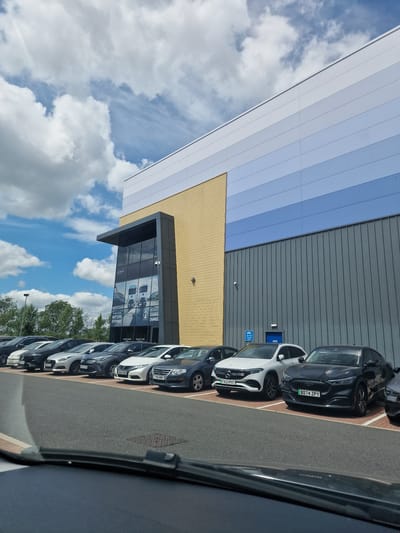 This is a photo of a row of cars parked in a lot outside a modern building with a grey corrugated metal facade and a section of yellow facade above a glass-fronted entrance. The building appears to be a business or showroom, with signs visible through the glass. The sky is blue with fluffy white clouds, suggesting it is daytime and the weather is fair. The cars are parked in marked bays, and some of them have license plates visible, indicating they are likely in the United Kingdom. The image is oriented vertically, with the building and cars appearing at an angle. Text visible on the building includes "NEXT LEVEL EFFICIENCY" and "CHARGE CHANGE", and license plates read "GF22 700", "AUG2 RZW", "FWO WMX", "FX23 PXT", and "B074 ZPY". The scene conveys a sense of commercial activity or a car dealership.