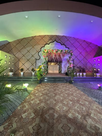 The image depicts an outdoor event venue, likely in Nashik, India, given the architectural style and the overall aesthetic. The venue features a decorated wedding stage with floral arrangements, set against a latticed wall backdrop illuminated with colored lights. The foreground shows patterned flooring leading to a stone-paved area with plants. The event appears to be set up for a celebration.