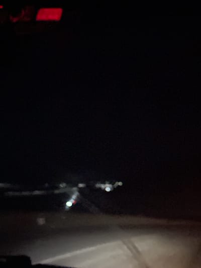 A blurry, dark image captures a view from inside a vehicle at night, likely in the vicinity of Беш-Мойнок, Kyrgyzstan. The scene is dominated by darkness, with some out-of-focus lights visible in the distance, suggesting a settlement or road infrastructure. The foreground shows a dimly lit road surface, possibly wet or covered in snow, with the glow of headlights creating a bright, hazy area. The top portion of the image reveals some red lights, which could be part of the vehicle's dashboard or external signage. There are no discernible people or specific structures identifiable due to the low light and lack of focus.