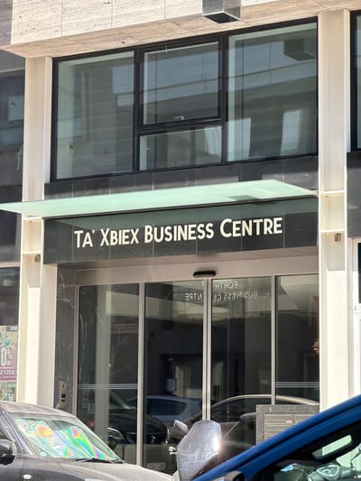 This is a medium-shot, eye-level, outdoor photograph of the entrance to the "TA' XBIEX BUSINESS CENTRE" in Ta' Xbiex, Malta. The building features a modern facade with dark granite or marble cladding and large glass windows. A frosted green glass awning extends over the entrance. White lettering on the dark cladding clearly reads "TA' XBIEX BUSINESS CENTRE". Below the awning, automatic glass doors lead into the building. Reflections in the glass doors show the mirrored text "BUSINESS CENTRE" and "HTROB" (likely a mirrored "FORTH"). In front of the entrance, a dark-colored car is parked, with its windshield covered by a reflective sunshade displaying rainbow patterns. Another car, predominantly blue, is visible in the foreground on the right, obscuring part of the scene. The overall lighting suggests a sunny day.