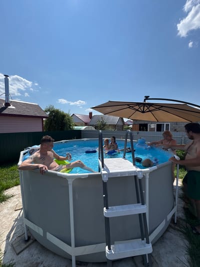 In a backyard in Uzuyukovo, Russia, a group of people are enjoying a day at an above-ground swimming pool. The pool is filled with blue water and surrounded by a gray liner. A white ladder with gray rungs provides access to the pool.

In the foreground, a man with tattoos on his chest and arm is sitting in the pool on a green inflatable ring, holding a can. To his right, a woman in a black swimsuit is leaning over the pool, holding a spray bottle, and interacting with a blonde-haired child submerged in the water. Beside her, another man in green shorts is standing and looking towards the woman.

In the background, several other individuals are in the pool, including two young girls and a child. A large, beige cantilever umbrella provides shade over part of the pool area. The backyard is grassy and features a wooden fence, houses with red roofs, and a chimney extending from one of the houses. The sky above is a clear blue with scattered white clouds.