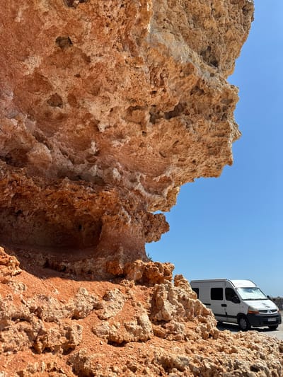 The image shows a close-up, upward angle view of porous, reddish-brown rock formations under a clear blue sky. The rock formations appear natural, with irregular shapes and numerous small holes and crevices. In the upper right corner, partially visible, is a white van parked on what appears to be a paved surface. The location is Balgarevo, Bulgaria.