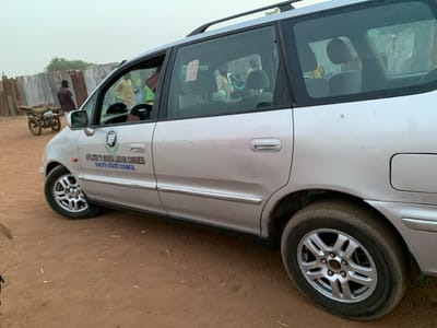 The image shows a silver minivan parked on a dirt road in Sokoto, Nigeria, located at coordinates 13.049 latitude and 5.238 longitude. The vehicle is facing to the right and has a large sticker on the side, reading "Affiliated to Nigeria Labour Congress Sokoto State Council." The vehicle is parked with its driver-side door open. There is a man in a blue and white shirt standing partially visible inside the vehicle. A second man in a red shirt is walking past the vehicle. There are more figures visible in the backseat through the rear windows, but they are blurred and not easily identifiable. The background includes a wooden fence and other unidentifiable objects. The ground is covered in dry, reddish-brown dirt. The sky is overcast.