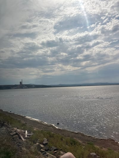 This media file depicts a scenic view of Port-la-Nouvelle, France, likely taken on a bright, overcast day. The image is oriented vertically, with the sky filling the left half and the sea the right.
The sky is characterized by dramatic, wispy clouds streaked with sunlight, suggesting a dynamic and possibly breezy weather pattern. The sun's rays create a bright lens flare on the left side of the frame.
The sea is a deep gray, with its surface shimmering and reflecting the sunlight, indicating a calm to moderately rough water condition. The shoreline, on the right side of the image, is a mixture of green grass and dark, rocky terrain. Some man-made debris, including white planks and stones, are visible along the embankment, hinting at human activity or past construction.
In the distance, across the water, a faint outline of the coastline is visible, with some industrial structures, possibly part of the port's infrastructure, breaking the horizon. These structures are a muted grey and blend into the atmospheric haze.
There are no visible people in the frame. The overall mood is one of natural beauty and the vastness of the seascape, with a subtle hint of industrial presence. The time of day appears to be late morning or early afternoon, given the brightness of the sun.