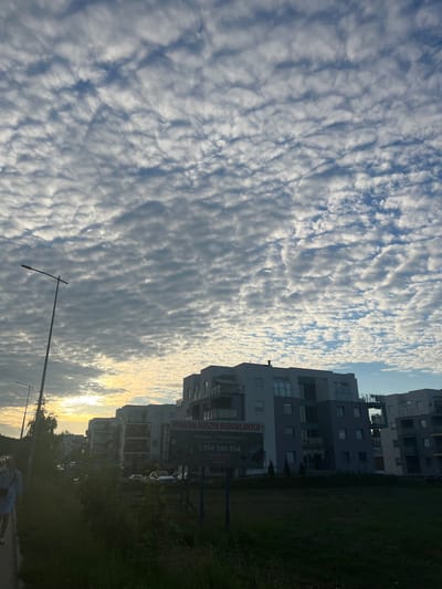 The image depicts a view of modern apartment buildings under a sky filled with altocumulus clouds. The sun is setting or rising, casting a warm glow on the horizon and illuminating the clouds. In the foreground, a street lamp stands to the left, and a person is walking on a sidewalk. A billboard is visible near the buildings, advertising "WYNAJEM MASZYN BUDOWLANYCH" (Rental of Construction Machinery) and providing a phone number. The location is identified as Rotmanka, Poland. The overall scene suggests a residential area during either sunrise or sunset.