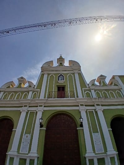 The image displays the ornate front facade of a church located in San Joaquín, Venezuela, captured from a low, upward-angle perspective. The building is painted predominantly in a pale green hue, accentuated by white architectural detailing, including columns, cornices, and decorative moldings.
The lower section of the facade features a large, central arched entryway fitted with dark brown double doors, characterized by recessed square panels. This central entrance is flanked symmetrically by two black wall-mounted lanterns. Two rectangular white plaques, likely commemorative, are affixed to the green wall below two smaller arched openings. Above the central entrance, a small balcony with a black metal railing precedes a red-brown door. A single arched window is positioned directly above this door, crowned by a white cross at the apex of the central gable.
Above the structure, multiple black utility lines are visible stretching across a clear blue sky. The sun is prominently featured in the upper right quadrant of the frame, producing a distinct lens flare effect and indicating a bright, clear day. No individuals or discernible activities are present within the scene.