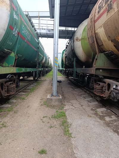 This media file depicts a railway yard with two rows of train tank cars, likely carrying industrial materials, parked on parallel tracks under a covered structure. The train car on the left is painted green and features the number "66" prominently, along with the sequence "51690063 21". The train car on the right appears older, with a weathered, yellowish-brown exterior showing signs of rust and peeling paint. It has visible markings including "51726". Both sets of train cars are situated on a railway track bed made of gravel and wooden sleepers, with some concrete pathways running alongside. The scene is outdoors, under a bright, overcast sky, suggesting daytime. There is some sparse vegetation growing between the tracks and along the edges of the concrete. The overall setting appears to be an industrial or transportation hub within Раснянскі сельскі Савет, Belarus. No people are visible, and the scene conveys a sense of stillness and industrial infrastructure.