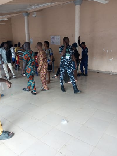 The scene depicts an indoor gathering within a light-brown walled hall featuring light-colored square tiled flooring and multiple white ceiling fans in Ajalli, Nigeria. At least ten individuals, primarily adults, are present. Two light-colored cylindrical columns support the ceiling. Several informational posters are affixed to the mid-ground wall.
In the mid-foreground, a male wears a black, grey, and white camouflage uniform with black combat boots and an identification badge on his chest; he is holding a dark mobile phone to his right ear, engaged in a conversation. To his left, two males are walking towards the right; one wears a teal, orange, and green patterned two-piece outfit with dark flip-flops, and the other wears an orange, red, and yellow patterned two-piece outfit with dark flip-flops. Further left, other individuals include a male with long dark hair wearing a striped shirt and light pants, and a female in a patterned dress. In the background-right, a male in a dark blue uniform with a red arm detail and dark cap stands with his left arm raised above his head, directed towards a column. Small pieces of debris, including a crumpled white object and a green object, are visible on the tiled floor.