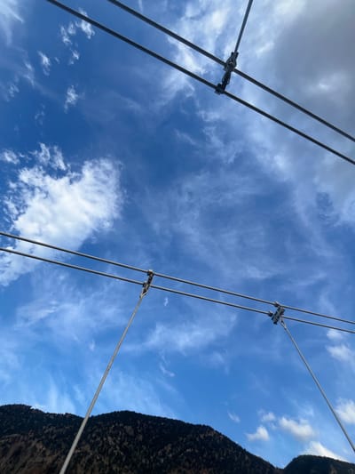 The image shows a view looking upwards at suspension cables against a bright blue sky with scattered white clouds. The cables are held together by metal clamps. The right edge of the frame includes a dark, textured mass, likely a rocky mountain face covered with vegetation. This scene is located in Ait Mazigh, Morocco.