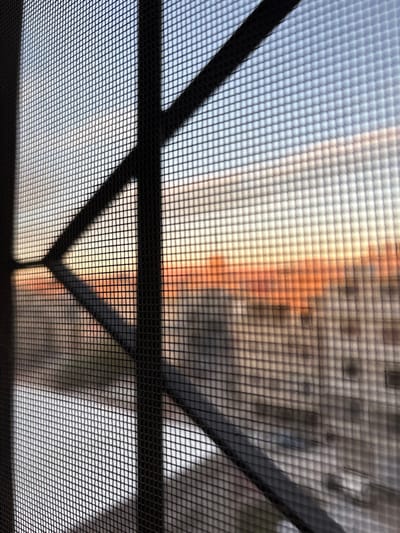 The image displays an urban landscape in Mu'adammiyat ash-Sham, Syria, viewed from an elevated perspective through a dark, finely meshed window screen and intersecting dark metal security bars. The screen and bars are in sharp focus in the foreground, creating a distinct grid pattern across the frame. The background is out of focus, featuring a sky that transitions from light blue at the top to warm orange and yellow hues near the horizon, consistent with a low sun position (sunrise or sunset). Below the sky, blurred forms of multi-story residential buildings are visible. No people, specific actions, or interactions are discernible within the scene.