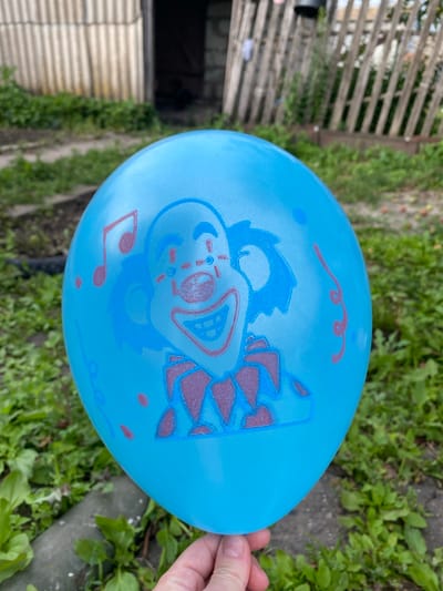 A close-up, slightly tilted, overhead shot shows a blue balloon held by a person's hand on the right. The balloon is printed with a cartoonish clown face in blue and red. Musical notes and squiggly lines are also printed on the balloon, suggesting a festive or celebratory theme. The background is blurred, revealing green grass and a wooden fence to the left, indicating an outdoor setting. The lighting suggests daytime, and the overall impression is casual and playful. No visible text is present on the balloon or in the background. The image is taken in Marinopil, Ukraine.
