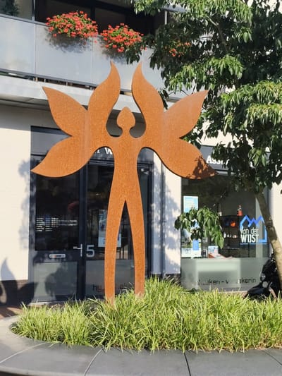 The image shows a contemporary outdoor metal sculpture in front of a building in Schaijk, Netherlands. The sculpture is of a stylized human figure with large, wing-like appendages extending from its shoulders. It is made of a reddish-brown, weathered metal, likely corten steel, and stands upright on a paved area. In the background, the building has large glass windows, through which signage for "Van der Wujst - hypotheken - verzekeringen - taxaties" is visible. The building also features a balcony with red flowers in planters and a tree with lush green foliage. The sculpture is surrounded by a bed of green, grass-like plants. The overall scene is brightly lit by sunlight, casting shadows on the ground and the sculpture. The image appears to be taken on a sunny day.