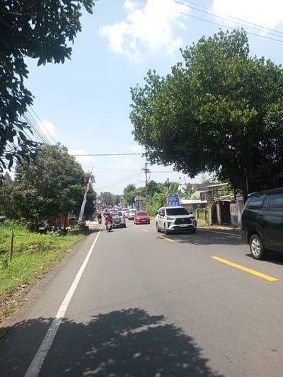 The image depicts a moderately busy road in an unspecified location that is likely not Nawng Hkar, Myanmar given contextual information. A mix of cars and motorcycles navigate the road, with a white car being most prominent in the center of the view. A black vehicle is partially visible on the right side of the image. The road is paved, with painted white and yellow lines to delineate lanes. Trees line both sides of the road, casting shadows on the road surface. The sky is blue with scattered clouds. Buildings and signage are visible in the background, suggesting a commercial or residential area. The image gives a sense of daytime traffic flow on a road in a populated area.