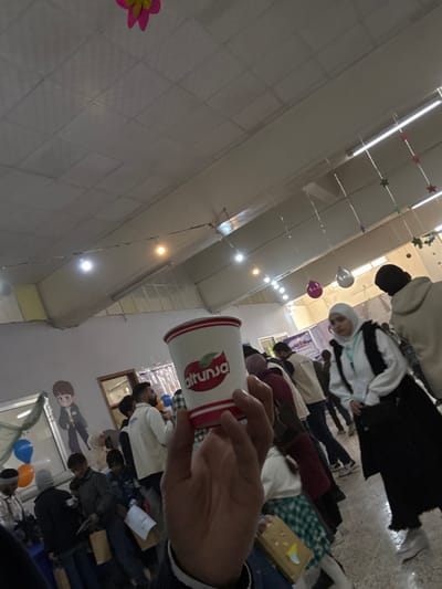This image depicts an indoor gathering in Kafr Batna, Syria. In the immediate foreground, a person's hand, wearing a dark-sleeved garment, holds a white paper cup with red horizontal stripes, a green and red apple logo, and the word "altunsa" printed in red text.
The background shows a hall populated by numerous individuals, comprising adults and some children, standing in various positions. The ceiling consists of light-colored, possibly acoustic, tiles and features exposed structural elements. Decorative string lights with bare bulbs are suspended across the ceiling, along with various colored balloons (purple, white, orange, blue) and star-shaped garlands.
In the mid-ground on the right, a female individual wears a white hijab, a white jacket over a dark top, a dark skirt, and a lanyard around her neck, carrying a dark shoulder bag. She is looking towards the left. Further left, a person is partially visible from behind, wearing a light-colored hoodie. Near the left background, a large cartoon-style cutout figure is displayed. Several other individuals, including a bearded man in a white long-sleeved shirt, are present. Display tables with items such as brown paper bags and a yellow box are visible in the background. A banner with Arabic text is partially discernible in the right background. The overall scene suggests a public event or exhibition.