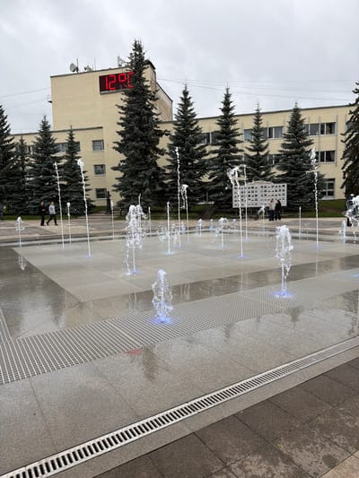 The image shows a city square in Belebei, Russia, with a modern fountain. The fountain features numerous jets of water, some illuminated with blue light, spraying upwards from a paved area. The paving stones are a mix of light grey and darker grey, with a metal drain running along one edge.

In the background, there is a light yellow building with several windows. On the side of this building, a large red digital clock displays the time "20:21". Evergreen trees, likely fir trees, are planted in front of the building and along the edge of the square, providing a contrast to the urban environment.

A few people are visible in the scene. Two individuals are standing near a display board which shows a grid of small photographs and some text. Another person is walking away from the viewer in the foreground, and a few more figures are further in the background, near the trees. The sky is overcast, suggesting a cloudy day. The overall impression is of a public space designed for recreation and gathering, with a blend of modern urban features and natural elements.