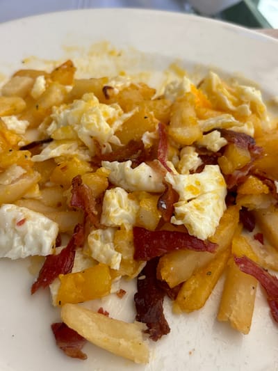 A plate features a medley of breakfast staples. Scrambled eggs, some still moist, are intermingled with golden-brown, roughly cubed fried potatoes. Strips of crispy bacon are scattered throughout, adding a savory element. The dish is presented on a white plate, with some areas appearing slightly greasy, suggesting a recent cooking.