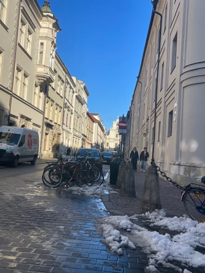 This image captures a sunlit street scene in Kraków, Poland, characterized by historic urban architecture. The street is lined with tall, light-colored buildings featuring classical elements like molding and numerous rectangular windows, rising towards a clear blue sky. On the left side, a white delivery van with an orange logo (partially visible text like "10" or "DO") is parked, and further down, the ornate roofline of a building with a small dome is visible.
The street surface is paved with patterned blocks or cobblestones, which appear wet and reflective in places. Along the right side of the street, remnants of dirty, melting snow are piled up, indicating a winter or early spring setting. Several bicycles are parked near the curb, alongside dark grey bollards connected by a chain. In the middle distance, a red and white "No Entry" sign is visible. Two individuals, dressed in winter jackets, are walking away from the camera on the right-hand sidewalk. The bright sunlight suggests it is daytime, casting clear shadows and contributing to a calm, ordinary urban atmosphere.