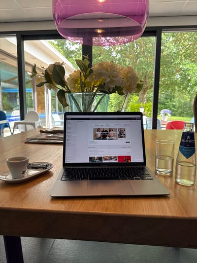 A MacBook Air laptop is open on a wooden table in front of a large window. The laptop screen displays the Shopify admin interface, with an online store theme and various options visible. To the left of the laptop, a white espresso cup sits on a saucer, next to a foil-wrapped biscuit. A clear glass of water and a bottle of Sourcy Naturel water are positioned to the right of the laptop. In the background, through the window, lush green trees and foliage are visible, suggesting an outdoor setting. A bouquet of white and yellow hydrangeas in a clear glass vase is placed on the table between the laptop and the window. The scene appears to be set in a modern, bright room with a prominent purple pendant light fixture hanging from the ceiling. The location is identified as Sint Maartensvlotbrug, Netherlands.