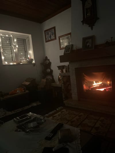 A dimly lit room features a fireplace with visible flames, and above it are framed pictures and a clock. A window, partially obscured by blinds and adorned with string lights, is also visible. A table covered with a white tablecloth is situated beside the fireplace, holding items like a remote control and a dish. The scene is dimly lit, creating a cozy and warm atmosphere.