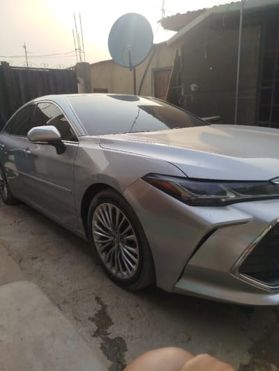 A silver Toyota Avalon is parked outdoors. The car's front passenger side is prominent, showcasing its modern headlight design and multi-spoke wheels. The background features a residential setting with a tall satellite dish, fencing, and a modest house. The lighting is somewhat subdued, possibly indicating early morning or late afternoon. Concrete surfaces surround the car, suggesting a driveway or yard.