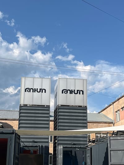The image shows a street scene in Gladzor, Armenia, with a bright blue sky and scattered clouds indicating daytime. The foreground features several shipping containers that have been repurposed. Two white containers are stacked vertically, each displaying the same black logo, which appears to read "runun". The logo is centered on the front of each container and oriented vertically. To the right of these, more grey shipping containers are visible, some forming part of a multi-story structure. Behind the containers, a building with a pinkish-brown facade and white-framed windows can be seen. Electrical wires crisscross the sky. The overall impression is one of urban redevelopment or a contemporary commercial space utilizing modular construction. No people are visible, and the scene appears to be static.