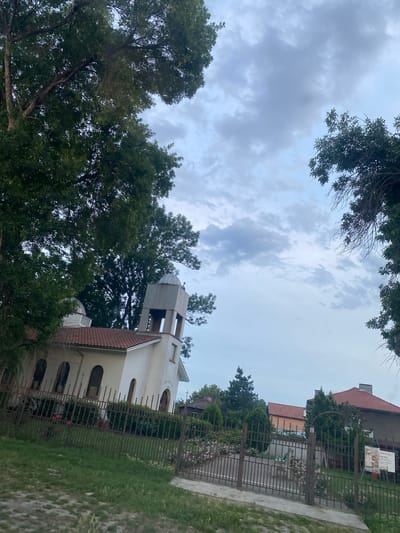 This is a vertical, eye-level shot of a white church with a bell tower, surrounded by trees and a fence. The sky is overcast with grey clouds. The church has arched windows and a red-tiled roof. A black metal fence runs along the front of the church, separating it from a grassy area and a path. To the right of the church, a building with a red roof and a sign is visible. The trees are lush and green, with some branches extending into the frame from the top and sides. The overall mood is peaceful and serene. The sign near the building is difficult to read but appears to be in a European language. The scene suggests it is daytime, likely morning or afternoon, given the lighting conditions. The location is Miriantsi, Bulgaria.