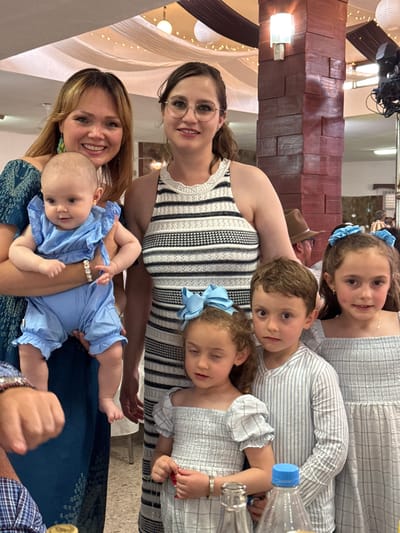 A group of seven people pose for a photograph indoors. From the bottom up, a woman with blonde hair smiles and holds a baby dressed in blue. Behind them, a woman with glasses smiles, and behind her are three children wearing blue and white striped clothing and blue bows. A water bottle is also visible. A man wearing a hat is partially visible behind the woman with glasses. The location appears to be a celebratory event, suggested by the decorations in the background, possibly in the city of Chipilo de Francisco Javier Mina (Chipilo), Mexico.