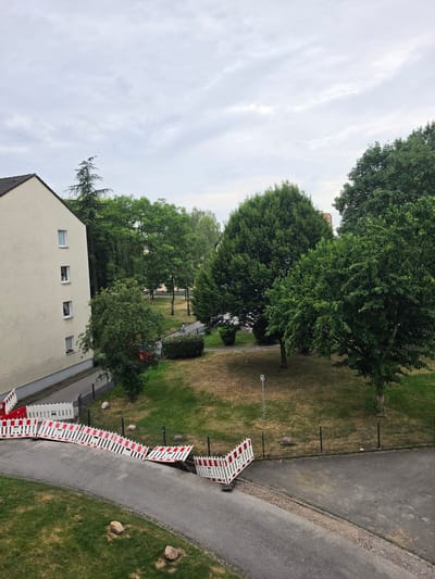 This is an outdoor, high-angle, full shot of a residential area in Oberhausen, Germany, under a cloudy sky. In the foreground, a paved pathway curves to the left, bordered by a grassy area with several large rocks. A temporary construction barrier, consisting of interlocking white and red panels, is set up across the pathway and the adjacent grassy area, with some sections appearing to be dislodged or collapsed.

To the left of the pathway, a light-colored apartment building runs vertically along the edge of the frame. It has several windows visible, some with occupants inside. Behind the building, lush green trees of various sizes and types create a dense backdrop.

Further into the scene, a park-like area with patchy dry grass and scattered trees is visible. A dark metal fence runs along the edge of this park area. In the middle ground, a person on a bicycle is seen on a paved path within the park, surrounded by trees. A tall building with a reddish-orange facade is visible in the distance, peeking out from behind the trees. The overall environment suggests an urban setting with a mix of residential buildings and green spaces.