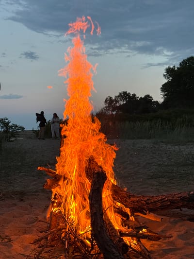 A bonfire burns on a sandy beach at dusk. The flames are high and bright orange, consuming several branches. Two people stand in the background, slightly obscured by the flames. The sky is a pale blue-gray, and there is vegetation along the shoreline in the background. The location is likely near Novo Oriahovo, Bulgaria, given the prompt's information.