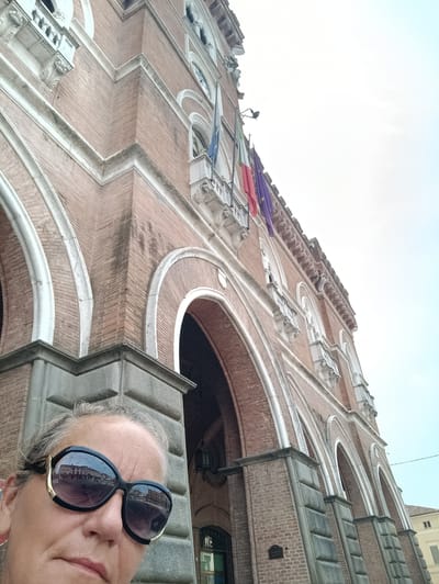 In a low-angle, upward shot, a woman wearing large, dark sunglasses with gold accents poses in the foreground. Her face, partially obscured by the sunglasses, is illuminated by daylight. The sunglasses reflect a street scene with buildings. Behind her, a grand, red brick building with arched doorways and decorative white trim dominates the background. Several flags, including the Italian flag and an EU flag, are visible hanging from the building. The architectural style suggests a historical public building. The image appears to have been taken outdoors on a clear day, possibly in Casalmaggiore, Italy, given the flags and architectural style often found in Italian public spaces.